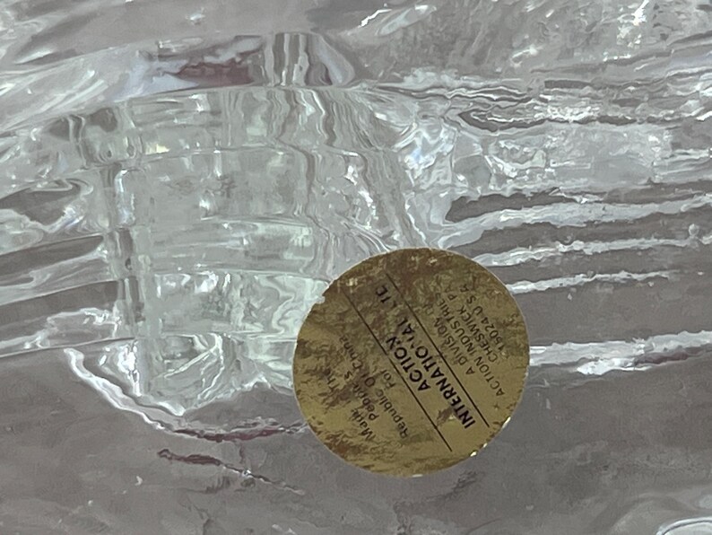 K&ouml;nnte beinhalten: Ein klares, strukturiertes Glasobjekt mit einem goldenen Aufkleber. Der Aufkleber tr&auml;gt die Aufschrift "ACTION INTERNATIONAL LIC" und enth&auml;lt Text &uuml;ber den Ursprung des Produkts in der Republik China.