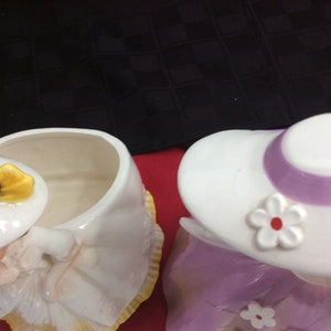 May include: Two ceramic cookie jars shaped like women wearing hats. The jar on the left is white with a yellow hat and bow. The jar on the right is purple with a white hat and a purple band. Both jars have a white flower on the hat.