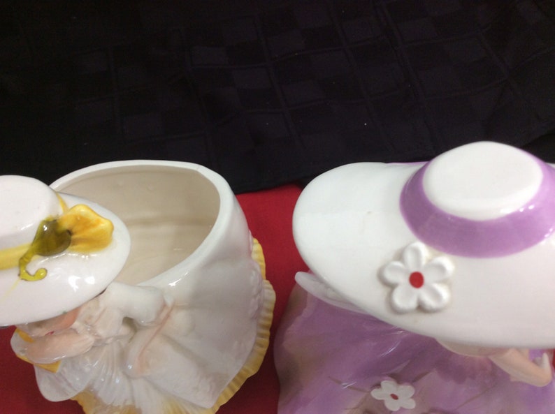 May include: Two ceramic cookie jars shaped like women wearing hats. The jar on the left is white with a yellow hat and bow. The jar on the right is purple with a white hat and a purple band. Both jars have a white flower on the hat.