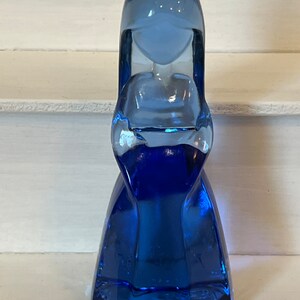 Pu&ograve; includere: Una statuetta in vetro blu cobalto a forma di figura stilizzata con testa arrotondata e abito fluente. Il vetro ha una superficie liscia e riflettente e la figura &egrave; in piedi. &Egrave; un oggetto decorativo.