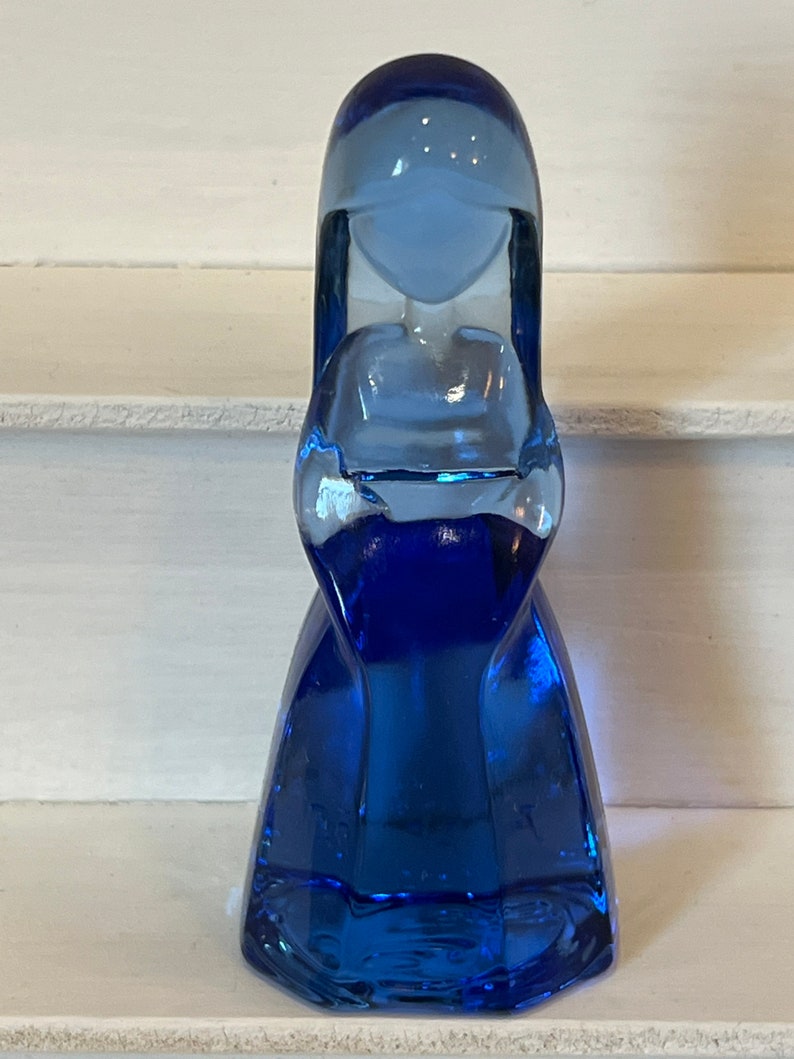 Pu&ograve; includere: Una statuetta in vetro blu cobalto a forma di figura stilizzata con testa arrotondata e abito fluente. Il vetro ha una superficie liscia e riflettente e la figura &egrave; in piedi. &Egrave; un oggetto decorativo.