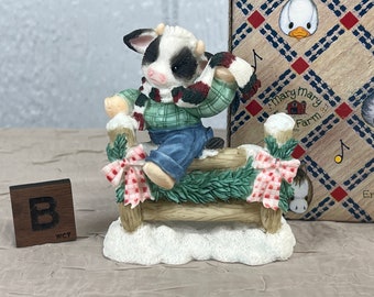 May include: A black and white cow figurine wearing a red and green plaid scarf and blue jeans, sitting on a wooden fence with a green garland and red and white bow. The figurine is part of a Mary Mary Farm collection.