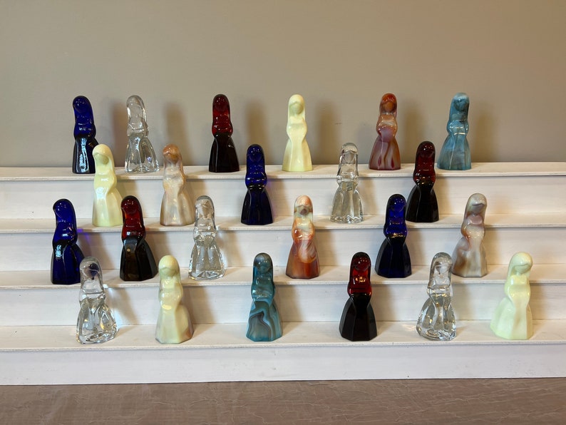 May include: A collection of 20 glass figurines arranged on a white tiered display stand. The figurines are all in the shape of a woman wearing a dress. The dresses are in various colors, including blue, red, white, and brown.