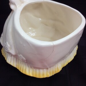 May include: White ceramic planter shaped like a woman in a dress with a yellow trim. The planter has a crack on the side.