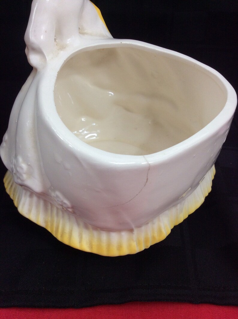 May include: White ceramic planter shaped like a woman in a dress with a yellow trim. The planter has a crack on the side.
