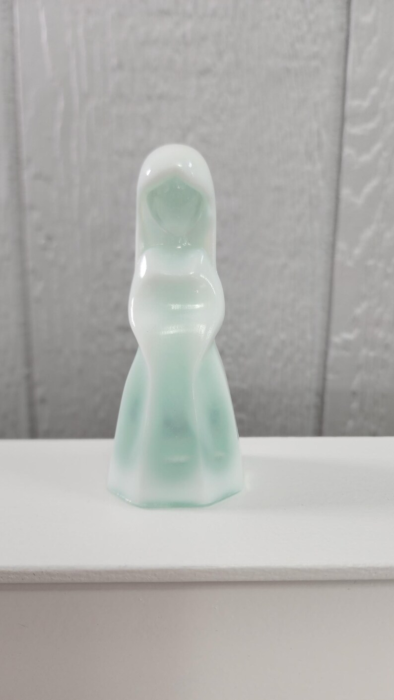 Pu&ograve; includere: Una statuetta in vetro traslucido, verde chiaro e bianco, raffigurante una figura incappucciata. La figura ha una testa arrotondata e una forma fluida, simile a un vestito. La statuetta &egrave; posta su una superficie bianca e uno sfondo grigio strutturato.