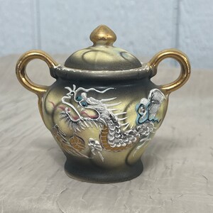 May include: A small, ceramic teapot with a gold lid and handles. The teapot is decorated with a white dragon and a butterfly on a black and yellow background.