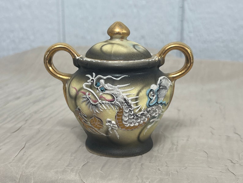 May include: A small, ceramic teapot with a gold lid and handles. The teapot is decorated with a white dragon and a butterfly on a black and yellow background.