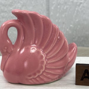 May include: A pink ceramic swan figurine with detailed wing feathers and a curved neck. The swan is set on a white surface, with a wooden block featuring the letter "A" and the initials "WCT".