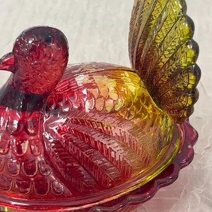 K&ouml;nnte beinhalten: Eine dekorative Glasschale in Form eines Fasans. Die Schale ist &uuml;berwiegend rot, mit gelben und gr&uuml;nen Akzenten. Die Schwanzfedern des Vogels sind leuchtend gelb-gr&uuml;n, und der K&ouml;rper hat eine strukturierte, irisierende Oberfl&auml;che. Sie dient wahrscheinlich zum Servieren oder Ausstellen.