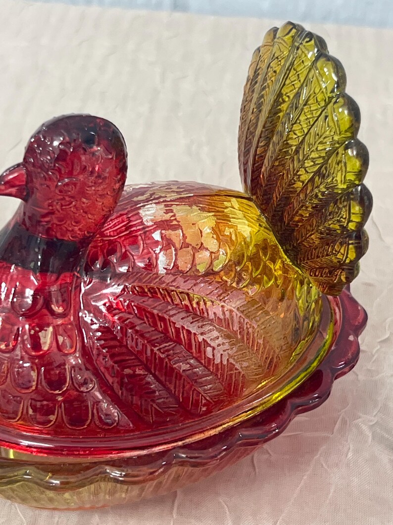 K&ouml;nnte beinhalten: Eine dekorative Glasschale in Form eines Fasans. Die Schale ist &uuml;berwiegend rot, mit gelben und gr&uuml;nen Akzenten. Die Schwanzfedern des Vogels sind leuchtend gelb-gr&uuml;n, und der K&ouml;rper hat eine strukturierte, irisierende Oberfl&auml;che. Sie dient wahrscheinlich zum Servieren oder Ausstellen.