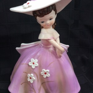 May include: A porcelain figurine of a girl wearing a white hat with a purple band and a purple dress with white flowers. The figurine is in a vintage style.