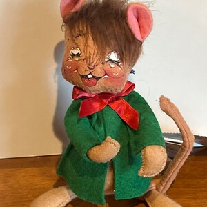 K&ouml;nnte beinhalten: Eine braune und beigefarbene Filz-Mauspuppe, die einen gr&uuml;nen Mantel mit einer roten Schleife tr&auml;gt. Die Maus hat braunes Haar, rosa Ohren und ein bemaltes Gesicht.