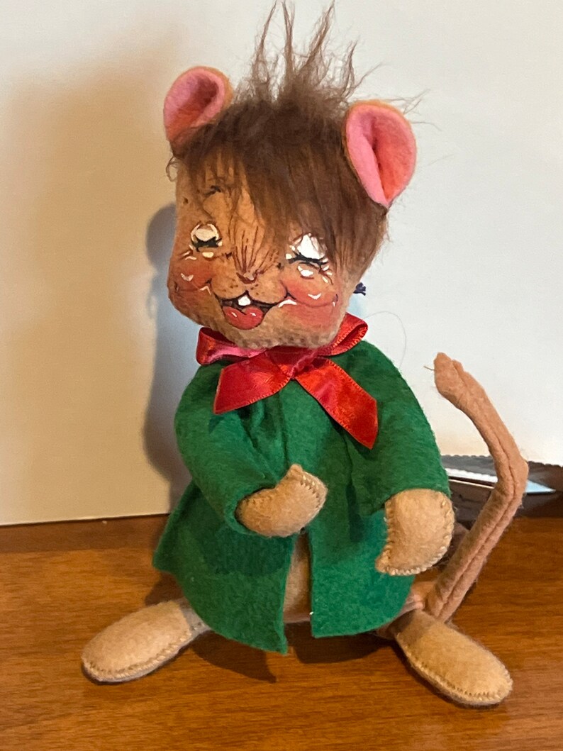K&ouml;nnte beinhalten: Eine braune und beigefarbene Filz-Mauspuppe, die einen gr&uuml;nen Mantel mit einer roten Schleife tr&auml;gt. Die Maus hat braunes Haar, rosa Ohren und ein bemaltes Gesicht.