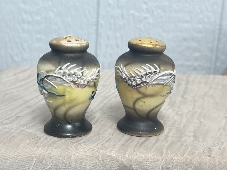 May include: A pair of ceramic salt and pepper shakers with a dragon design. The shakers are black, white, and yellow with gold accents.