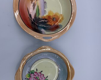 May include: Two vintage porcelain bowls with gold trim and painted floral and landscape designs. The bowls have two handles on each side.