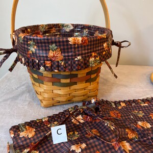 May include: A brown and tan wicker basket with a brown and white checked fabric liner featuring a fall leaf pattern. The basket has a wooden handle and a matching fabric liner with a fall leaf pattern. The liner has a ruffle around the top edge. The basket is sitting on a white surface. The liner has the letter "C" printed on it.