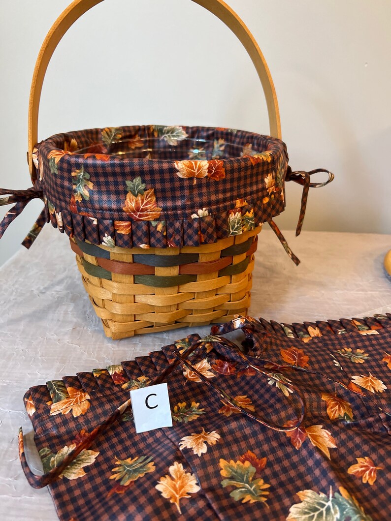 May include: A brown and tan wicker basket with a brown and white checked fabric liner featuring a fall leaf pattern. The basket has a wooden handle and a matching fabric liner with a fall leaf pattern. The liner has a ruffle around the top edge. The basket is sitting on a white surface. The liner has the letter "C" printed on it.