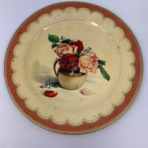 May include: A vintage tin plate with a red and gold border. The plate features a painted image of a bouquet of pink and red roses in a gold vase.