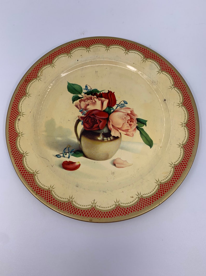 May include: A vintage tin plate with a red and gold border. The plate features a painted image of a bouquet of pink and red roses in a gold vase.