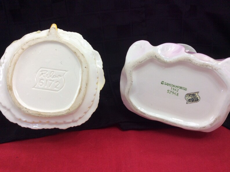 May include: Two vintage ceramic figurines, one white with a yellow floral design and the other pink with a floral design. The bottom of the white figurine has the text "Relco 5172" and the bottom of the pink figurine has the text "Samson Import Co. 1962 5296A".