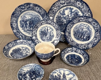 May include: A set of blue and white china dinnerware with a colonial theme. The plates feature scenes of colonial life, including a church, a town square, and a group of people riding horses. The set includes a large dinner plate, a salad plate, a soup bowl, a teacup, and a saucer.