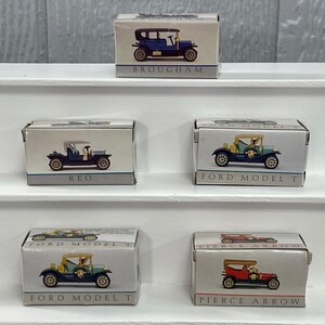 May include: Miniature car models in boxes, including a blue Brougham, a blue REO, and two Ford Model Ts in green and yellow. A red Pierce Arrow model is also visible. The boxes are white with the car names printed on them.