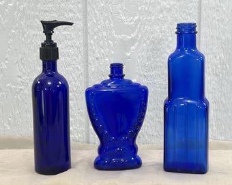Peut inclure: Trois bouteilles en verre bleu cobalt. L'une a un distributeur à pompe noir, une autre est de forme décorative et la troisième est de forme rectangulaire. Les bouteilles sont vides et ont une finition brillante. Elles sont probablement destinées à stocker des liquides.