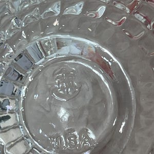 K&ouml;nnte beinhalten: Klares Glasobjekt mit einem strukturierten, gitterartigen Muster. Der Boden hat ein kreisf&ouml;rmiges Design mit dem gepr&auml;gten Text "USA". Das transparente Objekt l&auml;sst Licht durch und scheint ein Glaswarenst&uuml;ck zu sein.