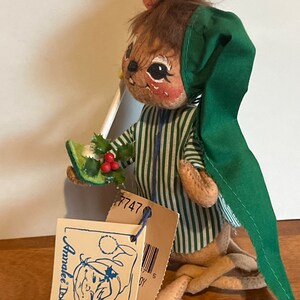 K&ouml;nnte beinhalten: Eine braune und gr&uuml;ne Filz-Mauspuppe, die einen gr&uuml;nen Hut und einen Pyjama tr&auml;gt. Die Puppe h&auml;lt einen gr&uuml;nen Stechpalmenzweig mit roten Beeren. Die Puppe hat ein Etikett mit der Aufschrift "Anne Geddes Dolls" und ein Preisschild mit der Nummer "7747".