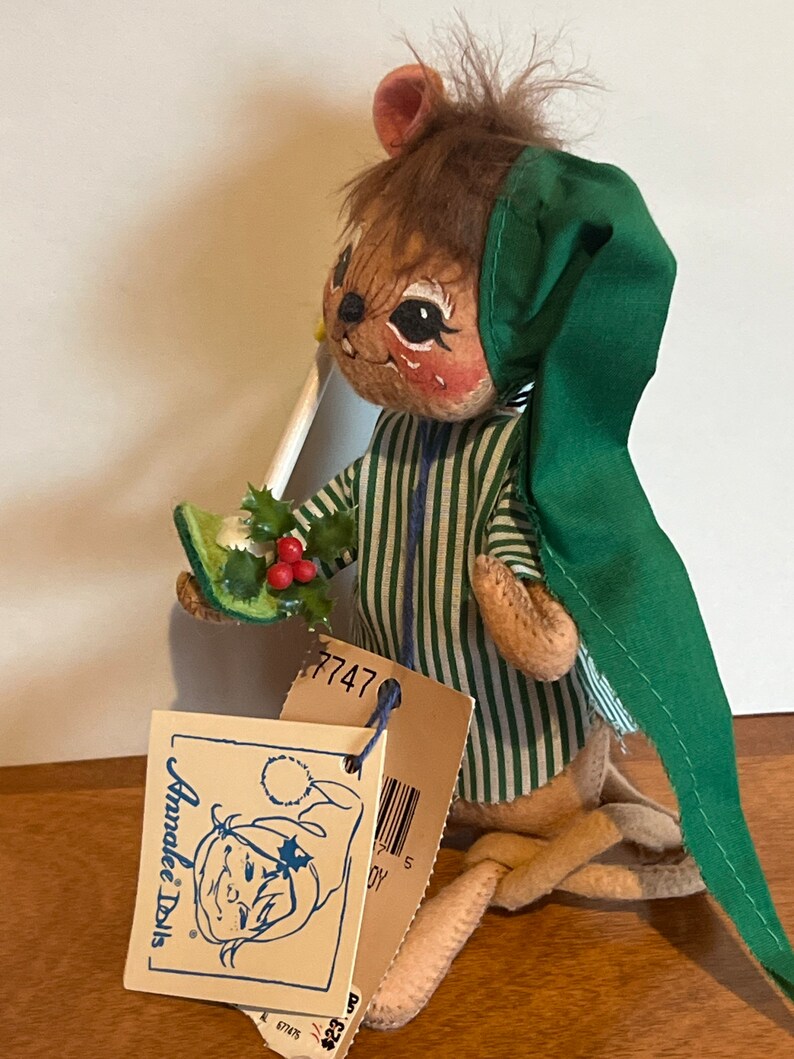 K&ouml;nnte beinhalten: Eine braune und gr&uuml;ne Filz-Mauspuppe, die einen gr&uuml;nen Hut und einen Pyjama tr&auml;gt. Die Puppe h&auml;lt einen gr&uuml;nen Stechpalmenzweig mit roten Beeren. Die Puppe hat ein Etikett mit der Aufschrift "Anne Geddes Dolls" und ein Preisschild mit der Nummer "7747".