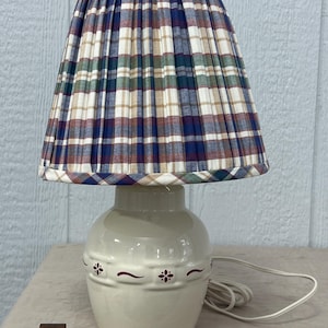 May include: A cream-colored ceramic table lamp with a plaid lampshade. The lampshade features a pleated design with a pattern of blue, green, brown, and white. The lamp base has a decorative floral design.