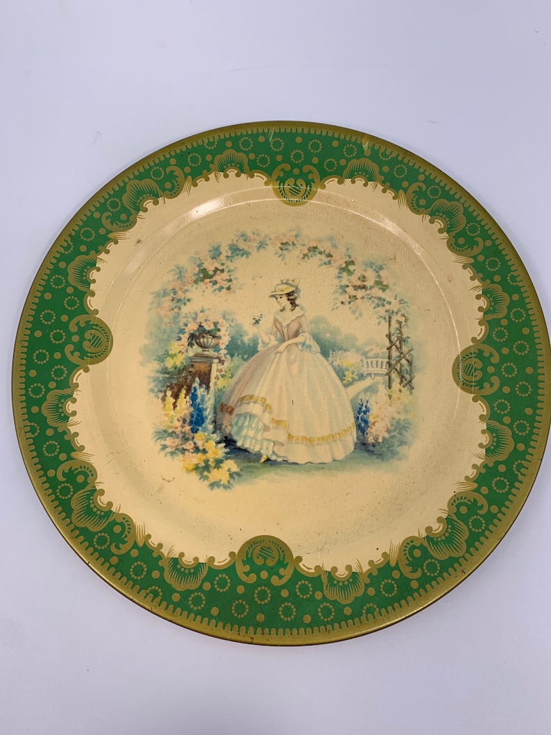 May include: A vintage metal serving tray with a green and gold border. The center of the tray features a painted image of a woman in a white dress standing in a garden.