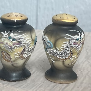 May include: A pair of ceramic salt and pepper shakers with a dragon design. The shakers are painted in shades of black, white, gold, and blue.