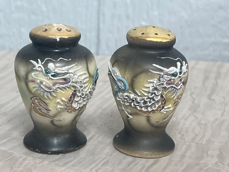 May include: A pair of ceramic salt and pepper shakers with a dragon design. The shakers are painted in shades of black, white, gold, and blue.