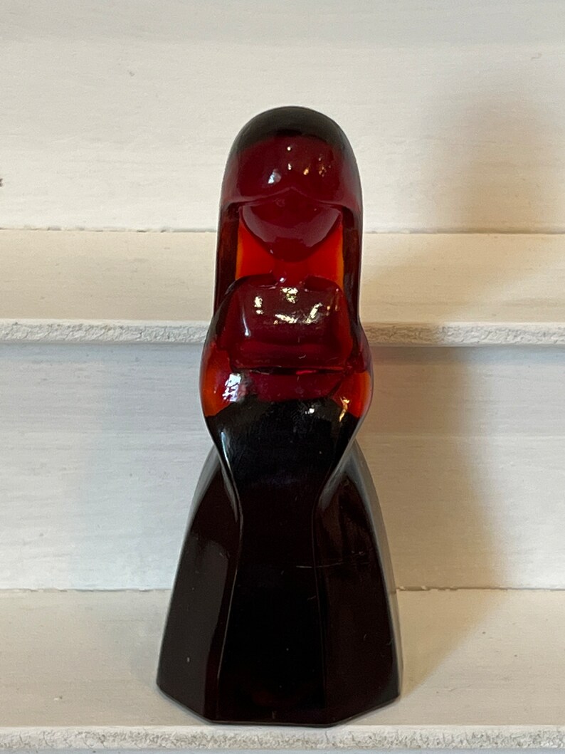 May include: A dark red glass figurine of a woman with long hair and a dress. The figurine is made of a dark red glass and has a glossy finish.