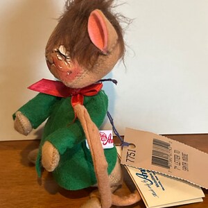 K&ouml;nnte beinhalten: Eine braune und beige Filzmaus, die ein gr&uuml;nes Kleid mit einer roten Schleife tr&auml;gt. Die Maus hat einen langen Schwanz und h&auml;lt ein Schild mit der Aufschrift "7" Brief an den Weihnachtsmann Maus".