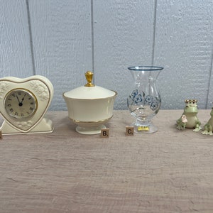 May include: Four decorative items arranged on a wooden surface. A heart-shaped clock with gold accents, a white porcelain lidded dish with gold trim, a clear glass vase with blue floral designs, and two green frog figurines wearing gold crowns.