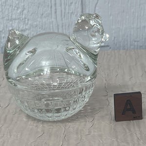 K&ouml;nnte beinhalten: Eine transparente Schmuckschatulle in Form eines Huhns mit strukturiertem Boden und Deckel. Das Huhn hat einen detaillierten Kopf und Fl&uuml;gel. Ein kleiner Holzblock mit dem Buchstaben "A" befindet sich neben der Box.