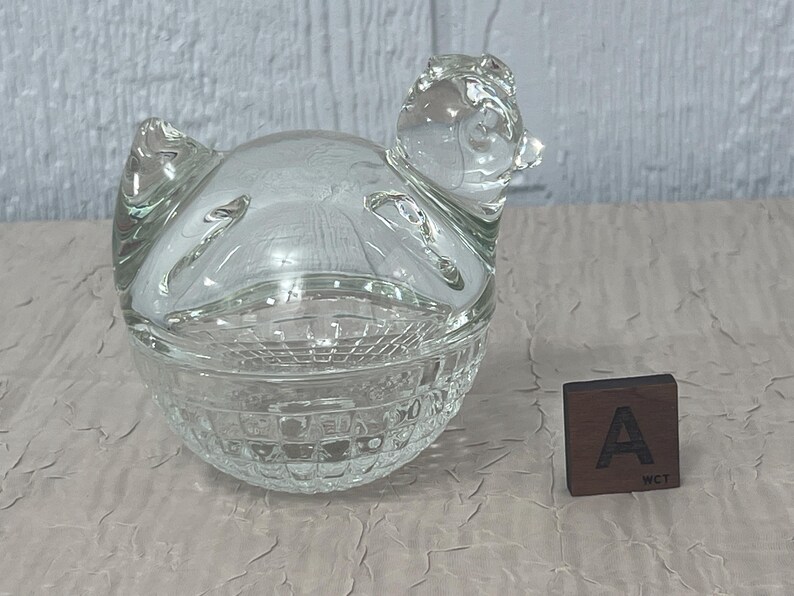 K&ouml;nnte beinhalten: Eine transparente Schmuckschatulle in Form eines Huhns mit strukturiertem Boden und Deckel. Das Huhn hat einen detaillierten Kopf und Fl&uuml;gel. Ein kleiner Holzblock mit dem Buchstaben "A" befindet sich neben der Box.