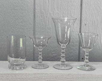 May include: Four clear glass drinking glasses of varying shapes and sizes are arranged in a row. The glasses include a tall tumbler, a stemmed coupe glass, a stemmed wine glass, and a small stemmed glass.