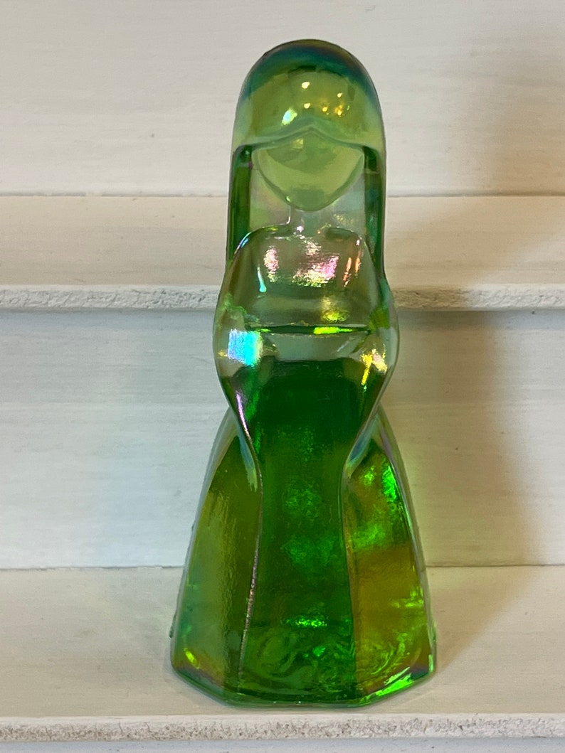 Pu&ograve; includere: Una statuetta in vetro verde iridescente raffigurante una donna in un lungo abito. La statuetta ha una lunga acconciatura e un'espressione serena. Il vetro ha un effetto cangiante multicolore, con sfumature di blu, viola e oro. La statuetta &egrave; alta circa 15 cm.