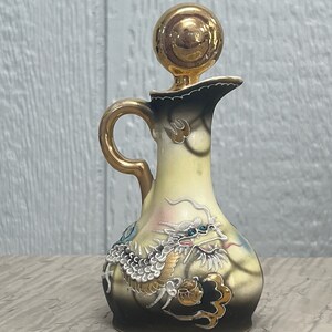 May include: A small, ceramic pitcher with a gold dragon design. The pitcher has a gold lid and a handle. The dragon is depicted in a yellow, white, and blue color scheme.