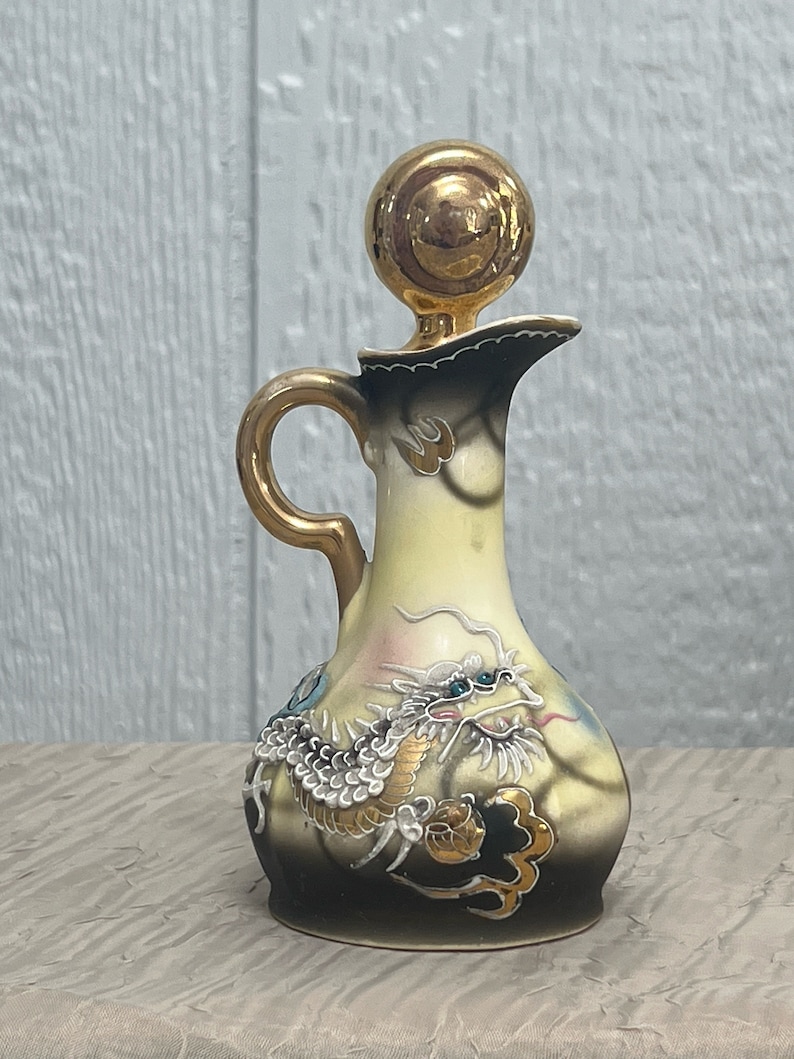 May include: A small, ceramic pitcher with a gold dragon design. The pitcher has a gold lid and a handle. The dragon is depicted in a yellow, white, and blue color scheme.