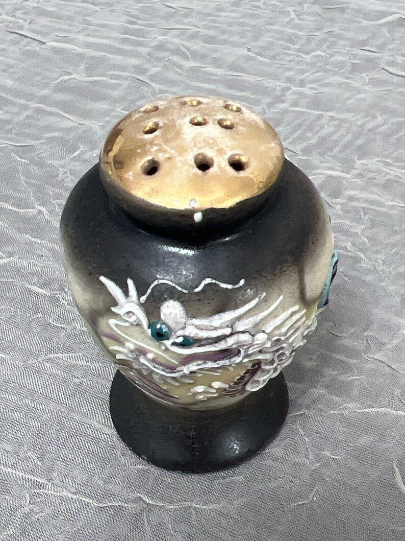 May include: A black ceramic salt shaker with a gold lid and a white dragon design. The lid has holes for dispensing salt.