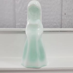 Pu&ograve; includere: Una statuetta decorativa bianca e verde chiaro a forma di figura incappucciata. La statuetta ha una finitura liscia e lucida e si trova su una superficie bianca. La forma della statuetta &egrave; semplice ed elegante.