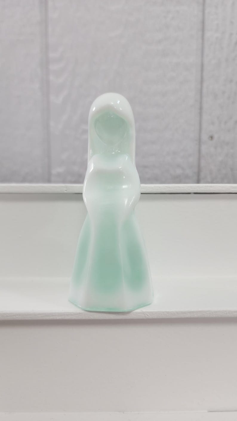 Pu&ograve; includere: Una statuetta decorativa bianca e verde chiaro a forma di figura incappucciata. La statuetta ha una finitura liscia e lucida e si trova su una superficie bianca. La forma della statuetta &egrave; semplice ed elegante.
