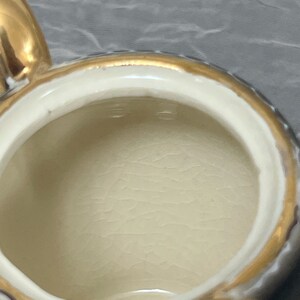 May include: A white ceramic teapot with a gold rim and handle. The inside of the teapot is visible and is white.