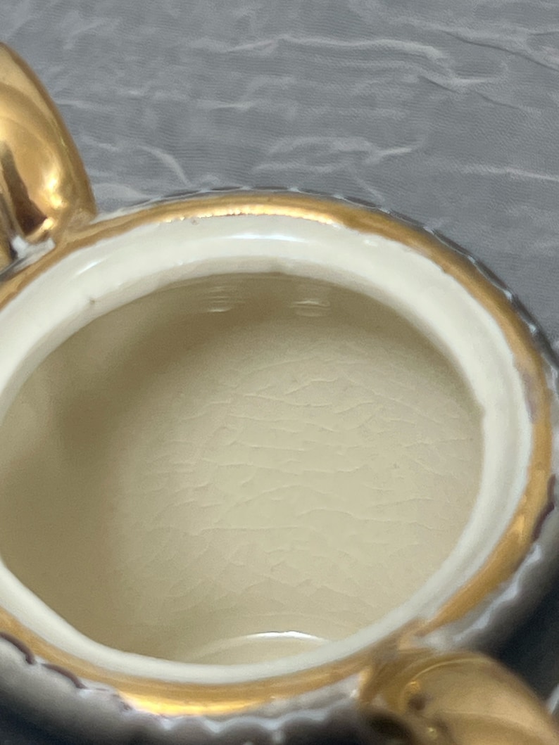 May include: A white ceramic teapot with a gold rim and handle. The inside of the teapot is visible and is white.