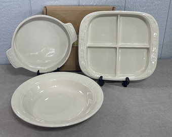 May include: A set of three off-white ceramic dishes including a round handled baking dish, a square four-section divided serving tray, and a wide shallow bowl. Each piece features a smooth, glossy finish and a subtle embossed border pattern.